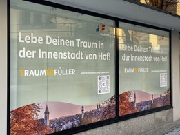 Das Bild zeigt ein Schaufenster, das mit einem Bild von Hof und der Aufschrift "Lebe Deinen Traum in der Innenstadt von Hof" beklebt ist.