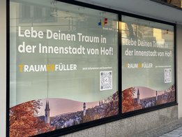 Das Bild zeigt ein Schaufenster, das mit einem Bild von Hof und der Aufschrift "Lebe Deinen Traum in der Innenstadt von Hof" beklebt ist.