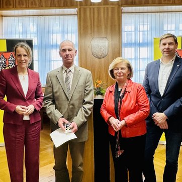 Auf dem Bild sind vier Personen in einem Saal zu sehen, hinter ihnen das Hofer Stadtwappen.