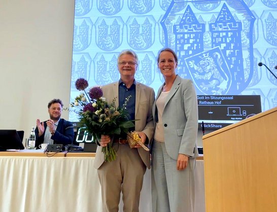 Auf dem Bild stehen ein Mann mit Blumenstrauß und eine Frau in einem Saal. Ein Mann hinter ihnen klatscht.