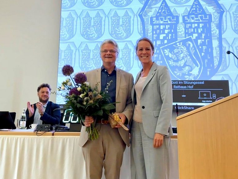 Auf dem Bild stehen ein Mann mit Blumenstrauß und eine Frau in einem Saal. Ein Mann hinter ihnen klatscht.