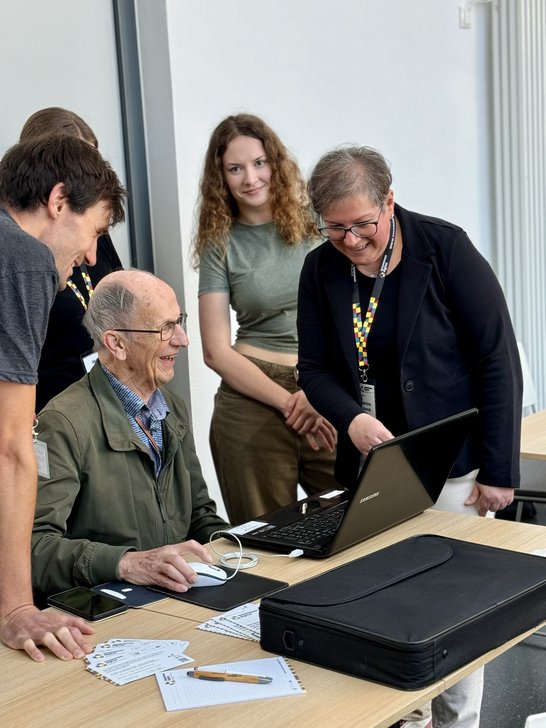 Auf dem Bild ist ein Senior zu sehen, der an einem Laptop sitzt. Um ihn herum stehen vier Personen.