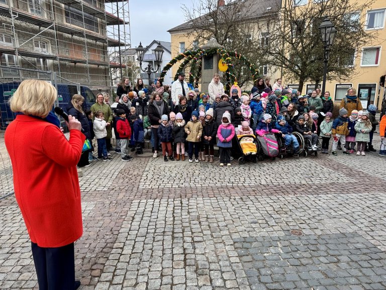 Auf dem Bild ist eine Frau zu sehen, die mit einem Mikrofon zu einer Menge Kinder spricht, die vor einem bunt geschmückten Brunnen stehen.