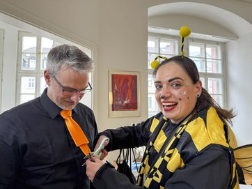 Auf dem Bild ist eine Frau im Bienenkostüm zu sehen, die einem Mann die Krawatte abschneidet.