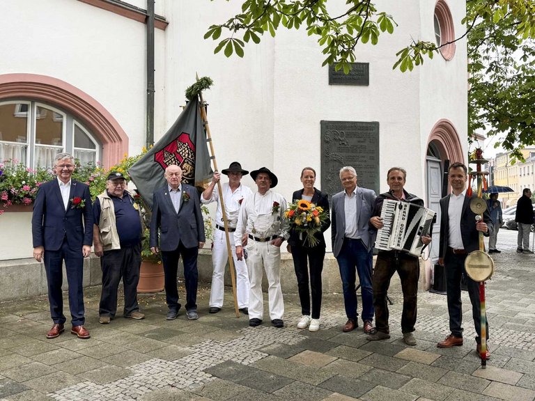 Auf dem Bild sind neun Personen zu sehen, davon zwei Handwerker in traditioneller Kleidung mit Fahne.