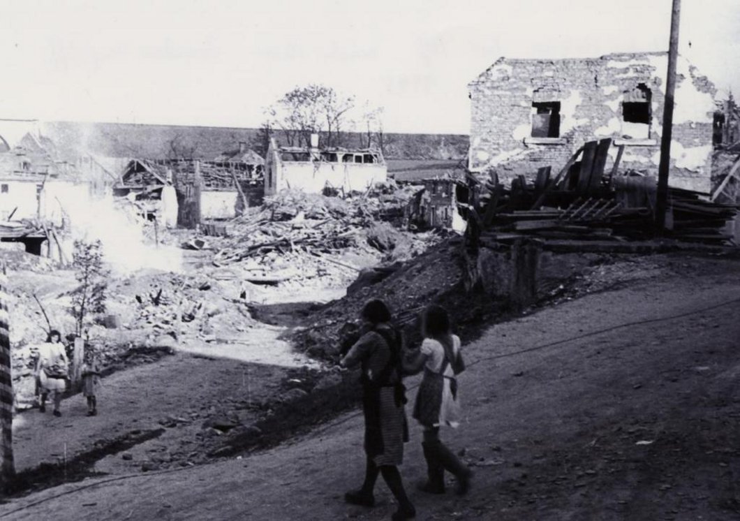 Auf dem Schwarz-Weiß-Foto ist eine völlig zerstörte Siedlung mit weggebombten Hausdächern zu sehen. Im Vordergrund gehen zwei Mädchen vorbei.