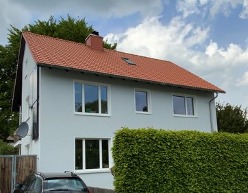 Auf dem Bild ist ein zweistöckiges Einfamilienhaus mit hellblauer Fassade und rotem Dach im Grünen zu sehen.