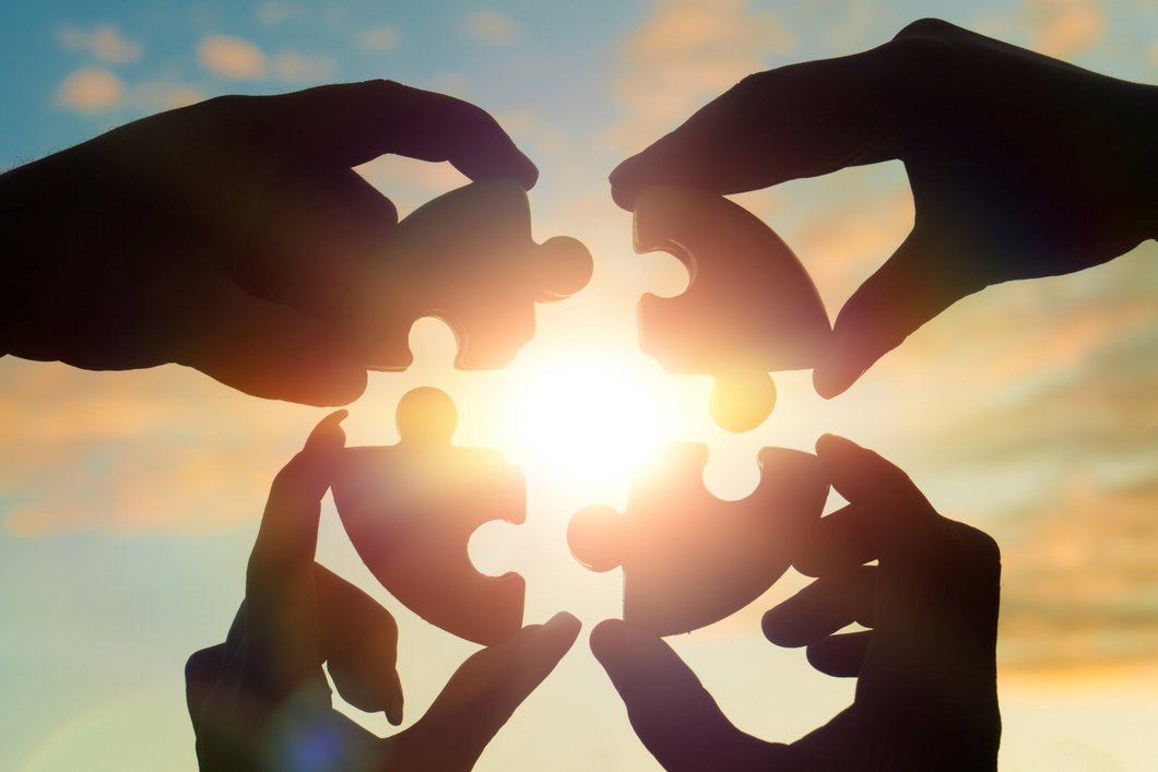 Auf dem Bild sind vier Hände vor der Sonne zu sehen, die Puzzleteile aneinanderhalten.