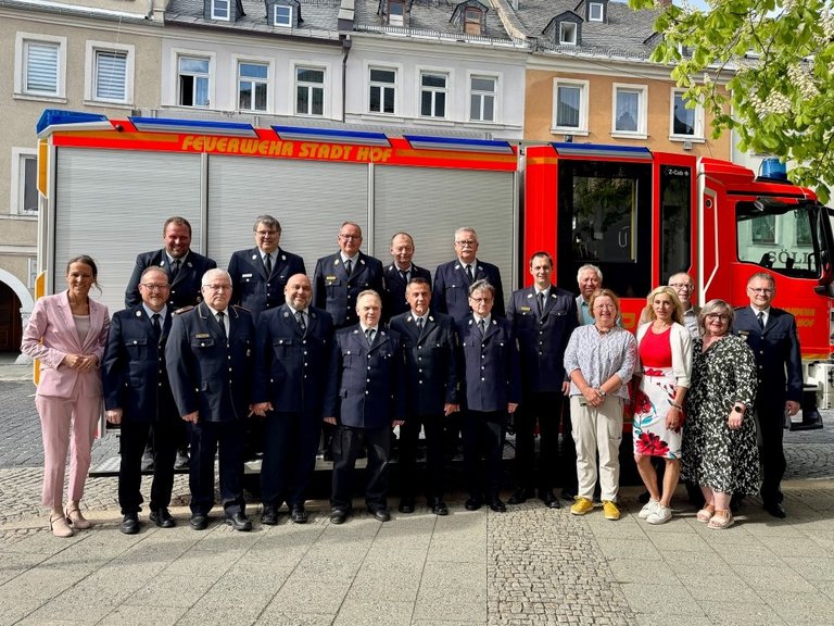 Auf dem Bild sind zwölf Feuerwehrmänner in Uniform und sieben weitere Personen vor einem Feuerwehrfahrzeug zu sehen.