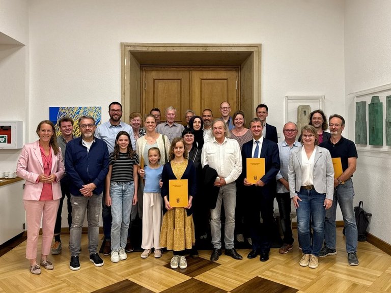 Auf dem Bild sind über 20 Personen vor einem Rathaussaale zu sehen, von den einige Urkunden halten.