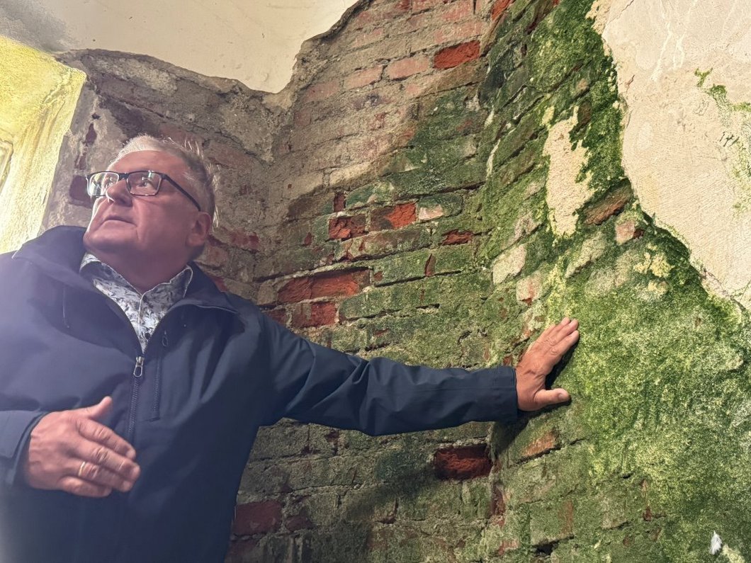 Auf dem Bild ist ein Mann zu sehen, der auf grün verfärbtes Mauerwerk zeigt, dessen Putz abgefallen ist.