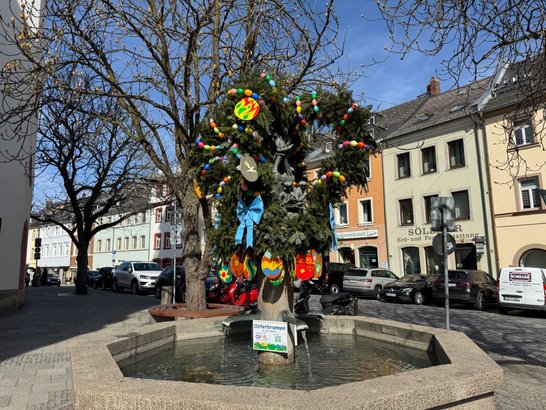 Auf dem Bild ist ein Brunnen mit grüner Krone zu sehen, die mit bunten Plastikeiern, bemalten Holzeiern und Schleifen geschmückt ist.