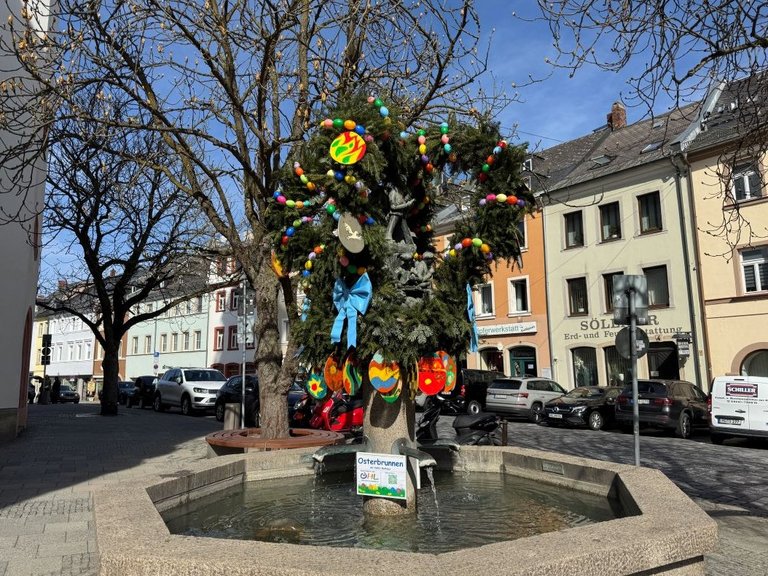 Auf dem Bild ist ein Brunnen mit grüner Krone zu sehen, die mit bunten Plastikeiern, bemalten Holzeiern und Schleifen geschmückt ist.