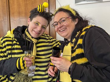 Auf dem Bild sind zwei Frauen im Bienenkostüm zu sehen, die miteinander anstoßen.