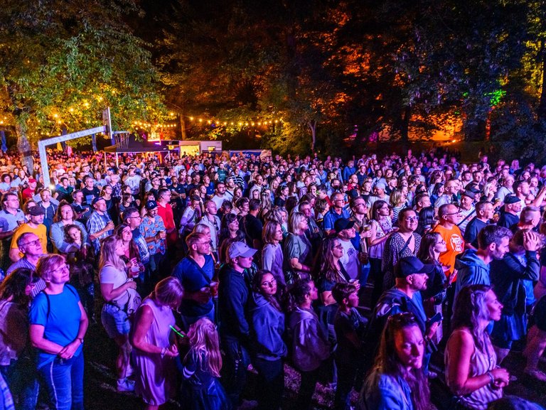 Auf dem Bild ist eine Menschenmenge am Abend auf einem cool beleuchteten Festivalgelände zu sehen.