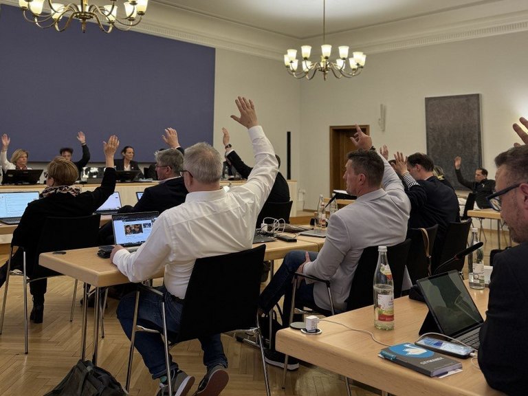 Auf dem Bild sind Personen in einem Sitzungssaal zu sehen, die die Hände zu einer Abstimmung heben.