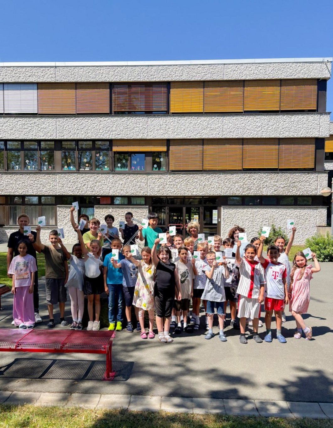 Auf dem Bild sind vier Erwachsene mit einer Schulklasse zu sehen, die vor einer Schule stehen. Die Kinder halten Flyer hoch.