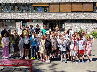 Auf dem Bild sind vier Erwachsene mit einer Schulklasse zu sehen, die vor einer Schule stehen. Die Kinder halten Flyer hoch.