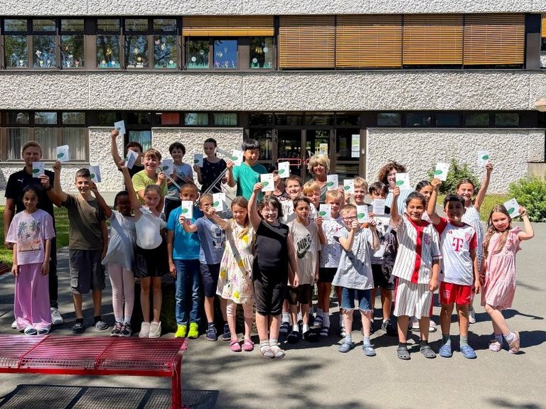 Auf dem Bild sind vier Erwachsene mit einer Schulklasse zu sehen, die vor einer Schule stehen. Die Kinder halten Flyer hoch.