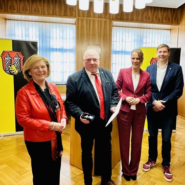 Auf dem Bild sind vier Personen in einem Saal zu sehen, hinter ihnen das Hofer Stadtwappen.