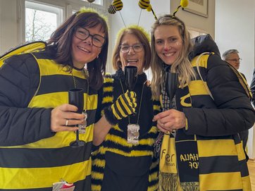 Auf dem Bild sind drei Frauen im Bienenkostüm zu sehen, die miteinander anstoßen. 
