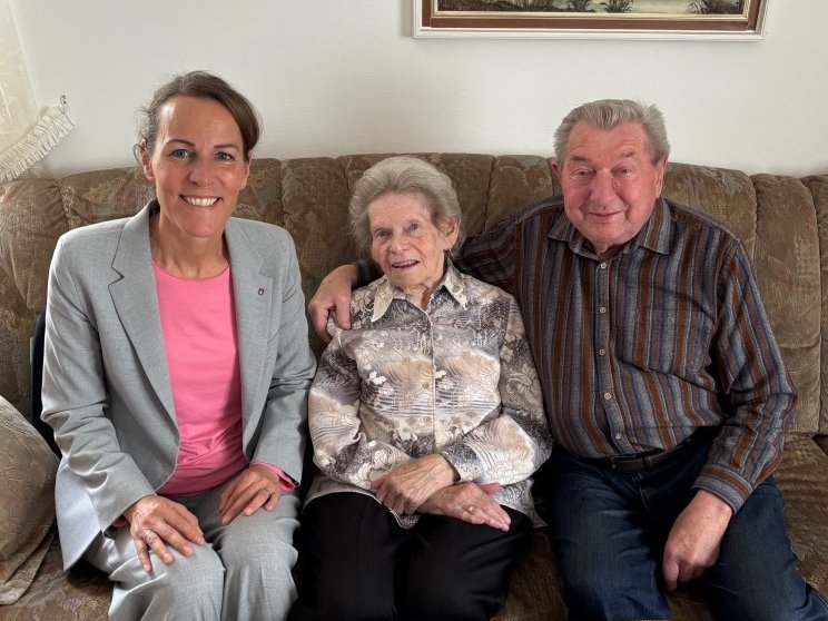 Auf dem Bild ist ein altes Ehepaar zu sehen, das mit einer Frau auf einem Wohnzimmersofa sitzt.