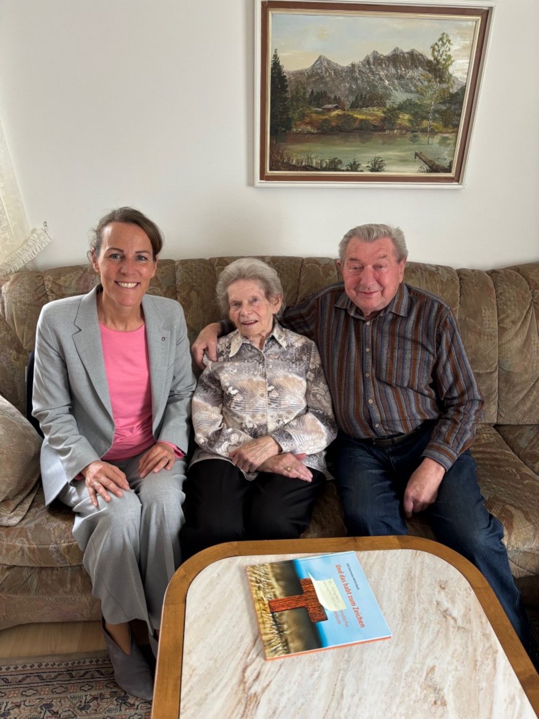 Auf dem Bild ist ein altes Ehepaar zu sehen, das mit einer Frau auf einem Wohnzimmersofa sitzt.