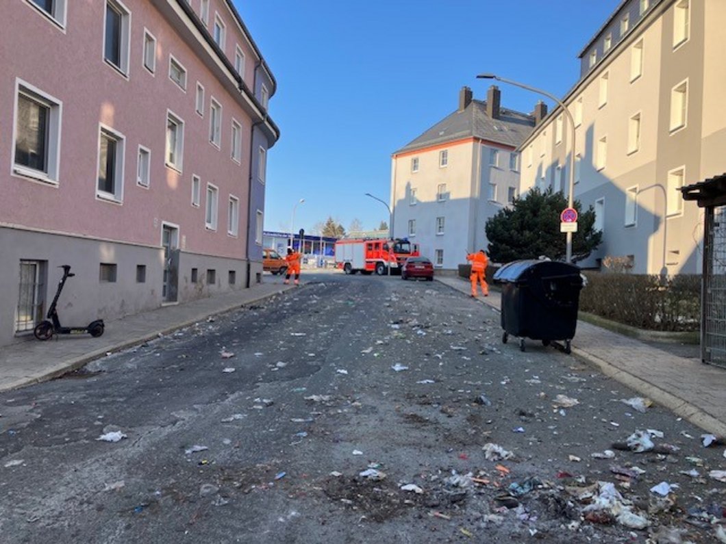 Auf dem Bild ist eine mit Müll überhäufte Straße zu sehen. Im Hintergrund abeiten orange gekleidete Männer.