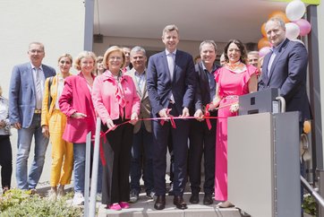 Auf dem Bild sind rund 15 Personen zu sehen, die vor einem Eingang stehen. Ein Mann schneidet ein rotes Band durch.