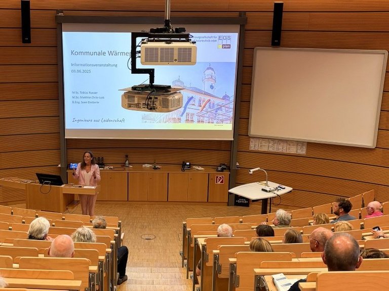 Auf dem Bild ist ein Vorlesungssaal mit Zuhörern zu sehen, die einer Rednerin zuhören.