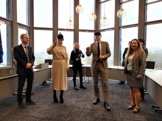 Auf dem Bild sind fünf Personen zu sehen, die in einem Sitzungssaal stehen. Eine Frau und ein Mann in der Mitte haben VR-Brillen auf und greifen mit der Hand in die Luft.