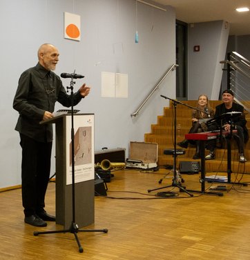 Auf dem Bild ist ein Mann zu sehen, der an einem Rednerpult steht. Im Hintergrund sitzen zwei Musiker.