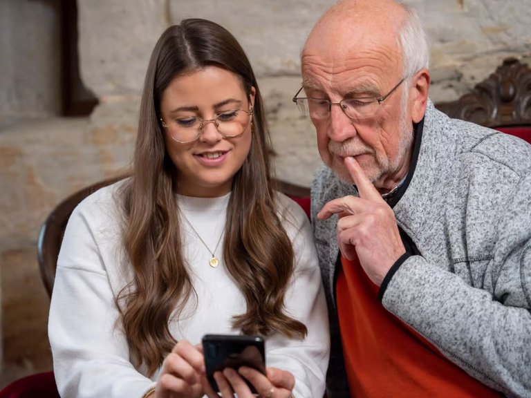 Dieses Bild zeigt eine junge Frau und einen alten Mann, die zusammen auf ein Smartphone schauen.