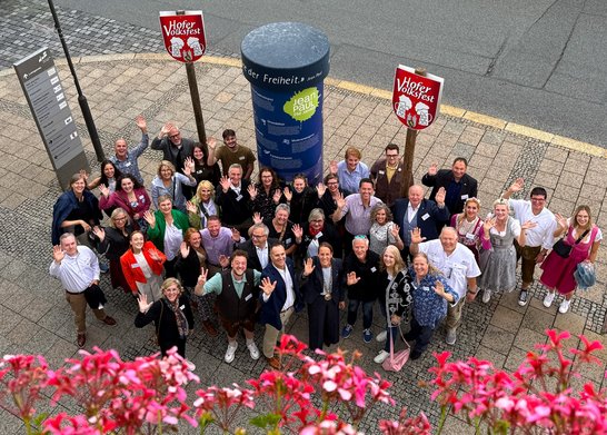 Auf dem Bild sind mehr als 30 Personen zu sehen, die zwischen Volksfestschildern stehen und winken.