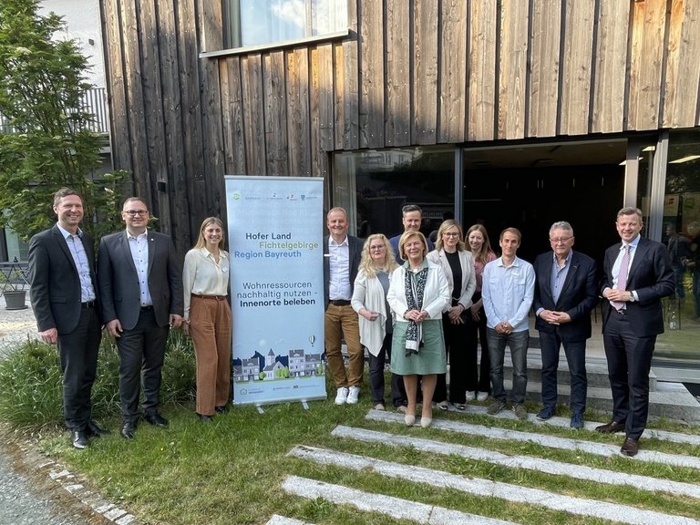 Auf dem Bild sind 12 Personen vor einem Holzgebäude zu sehen neben einem Aufsteller "Wohnressourcen nachhaltig nutzen)
