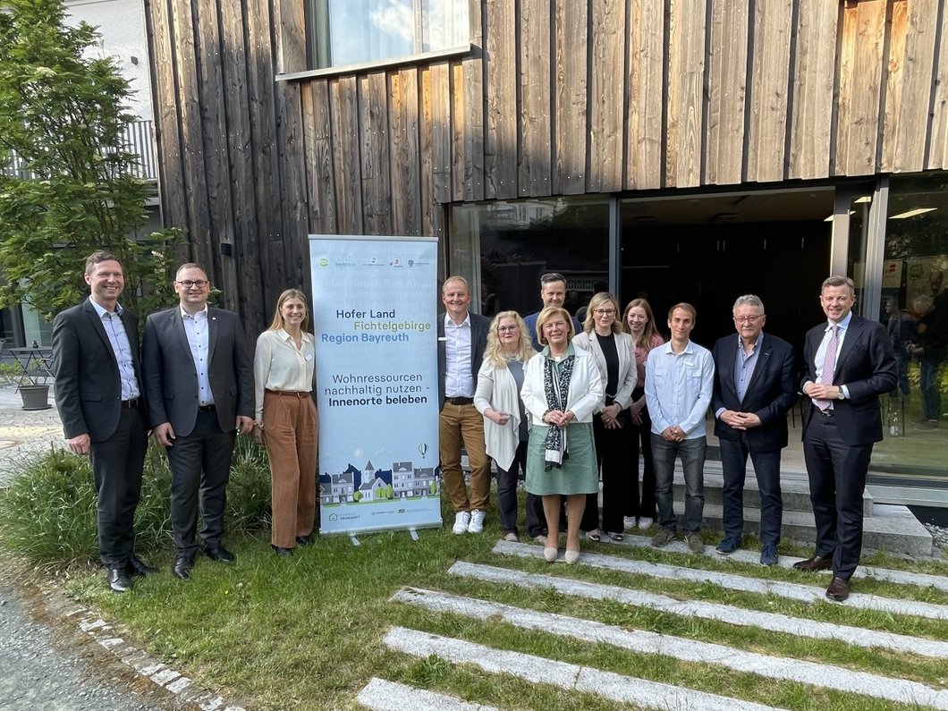 Auf dem Bild sind 12 Personen vor einem Holzgebäude zu sehen neben einem Aufsteller "Wohnressourcen nachhaltig nutzen)