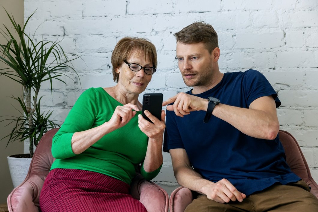 Auf dem Bild ist ein junger Mann zu sehen, der einer älteren Frau etwas an ihrem Smartphone erklärt.