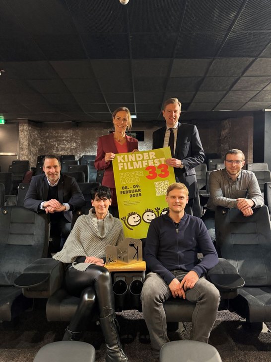 Auf dem Bild sind sechs Personen zu sehen, die sich in einem Kinosaal befinden. Zwei von ihnen halten ein Plakat hoch.