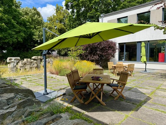 Auf dem Bild sind ein Holztisch mit Büchern und Stühlen zu sehen, die in unter einem Sonnenschirm stehen.