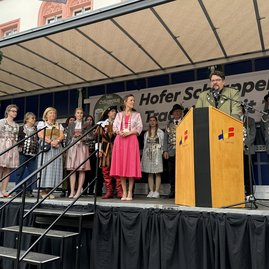 Männer und Frauen in Dirndln und Schützenuniformen stehen auf einer Bühne. Ein Mann in Schützenuniform steht an einem Pult und spricht.