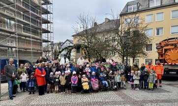 Auf dem Bild ist eine Menge von Kindern zu sehen, die vor einem bunt geschmückten Brunnen stehen. Daneben orange gekleidete Männer und ein oranges Fahrzeug.
