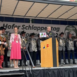 Das Bild zeigt einen Mann, der auf einer Bühne am Rednerpult steht und zwei bunte Schlappen aus Papier hochhält.