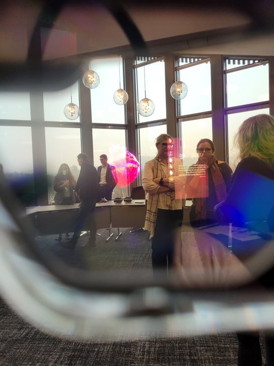 Das Bild ist durch eine VR-Brille aufgenommen. Im Hintergrund sieht man Menschen in einem Raum stehen, davor wird ein virtuelles Bild von einem Herze und ein Infokasten in den Raum projiziert.