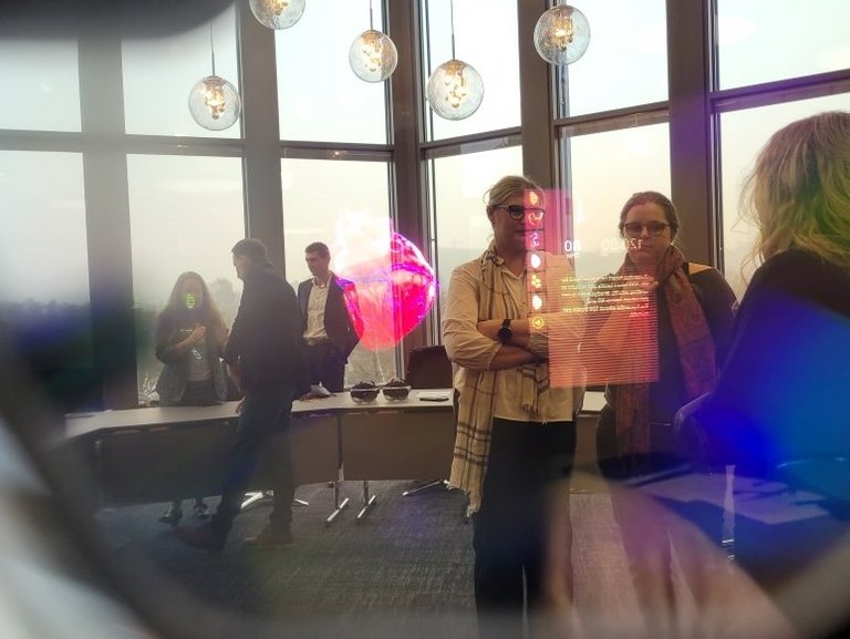 Das Bild ist durch eine VR-Brille aufgenommen. Im Hintergrund sieht man Menschen in einem Raum stehen, davor wird ein virtuelles Bild von einem Herze und ein Infokasten in den Raum projiziert.