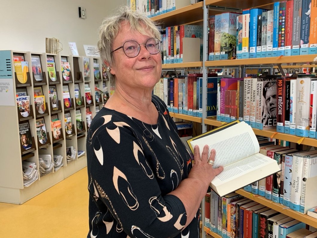 Auf dem Bild ist eine Frau zwischen Bücherregalen zu sehen, die ein offenes Buch hält.