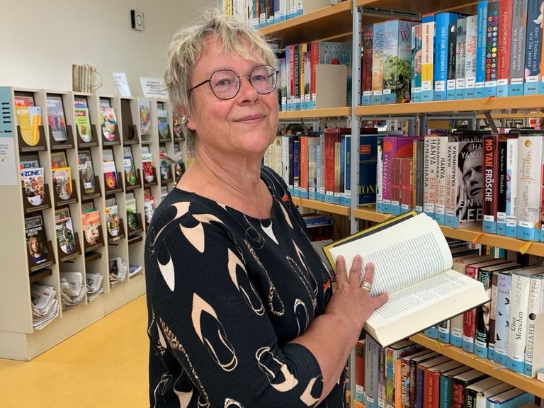 Auf dem Bild ist eine Frau zwischen Bücherregalen zu sehen, die ein offenes Buch hält.