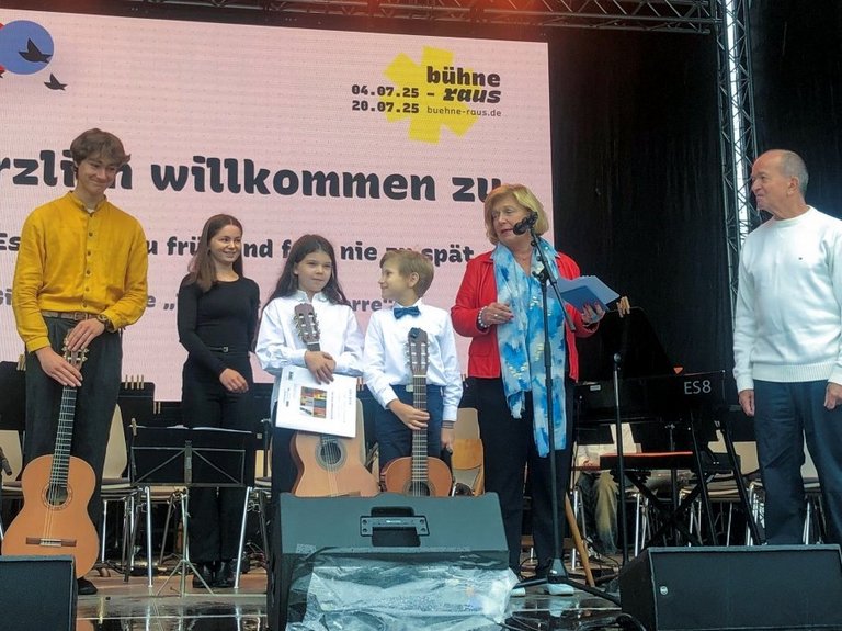 Auf dem Bild sind zwei Erwachsene zu sehen, die mit vier Kindern bzw. Jugendlichen auf einer Bühne stehen.
