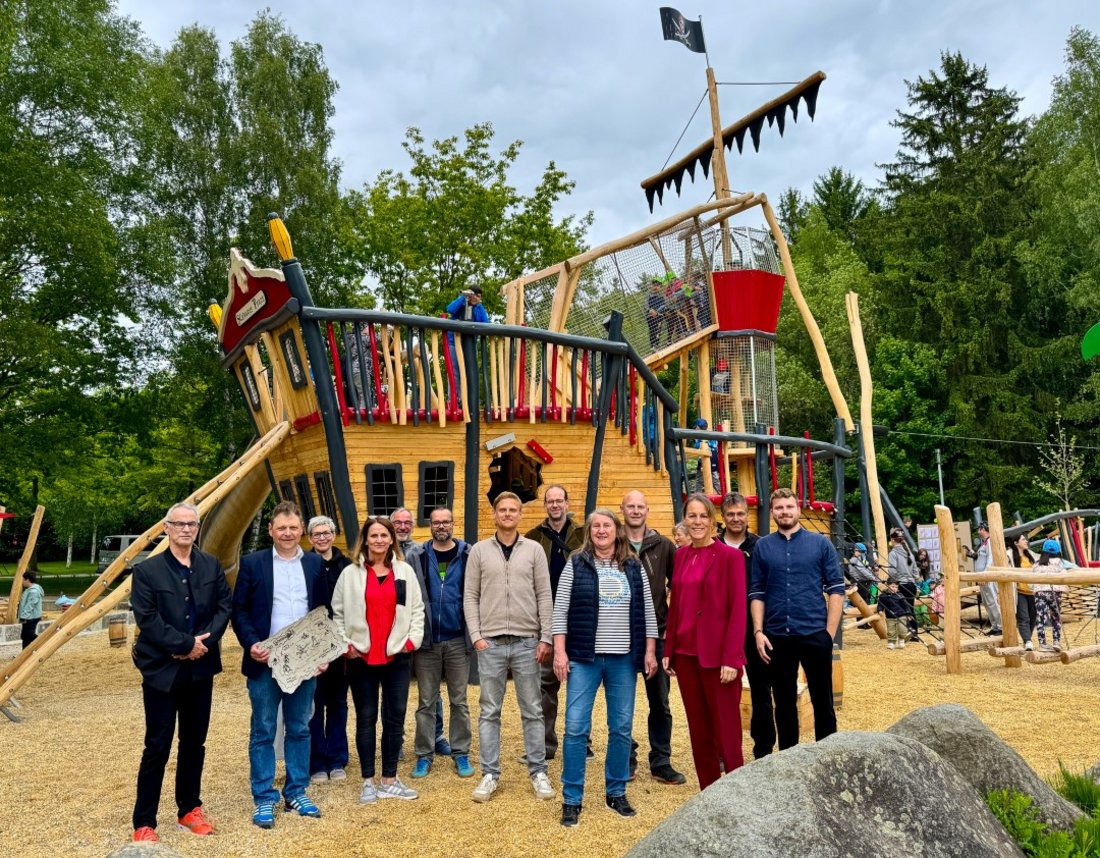 Auf dem Bild sind 13 Personen zu sehen, die auf einem Spielplatz stehen. Hinter ihnen ein hölzernes Piratenschiff.