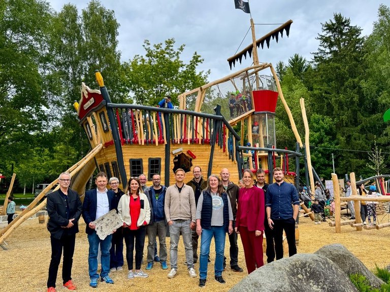 Auf dem Bild sind 13 Personen zu sehen, die auf einem Spielplatz stehen. Hinter ihnen ein hölzernes Piratenschiff.