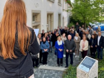 Auf dem Foto ist eine Frau von hinten zu sehen, die zu einer Menschenmenge spricht. Neben ihr steht eine Stele mit Infotext.